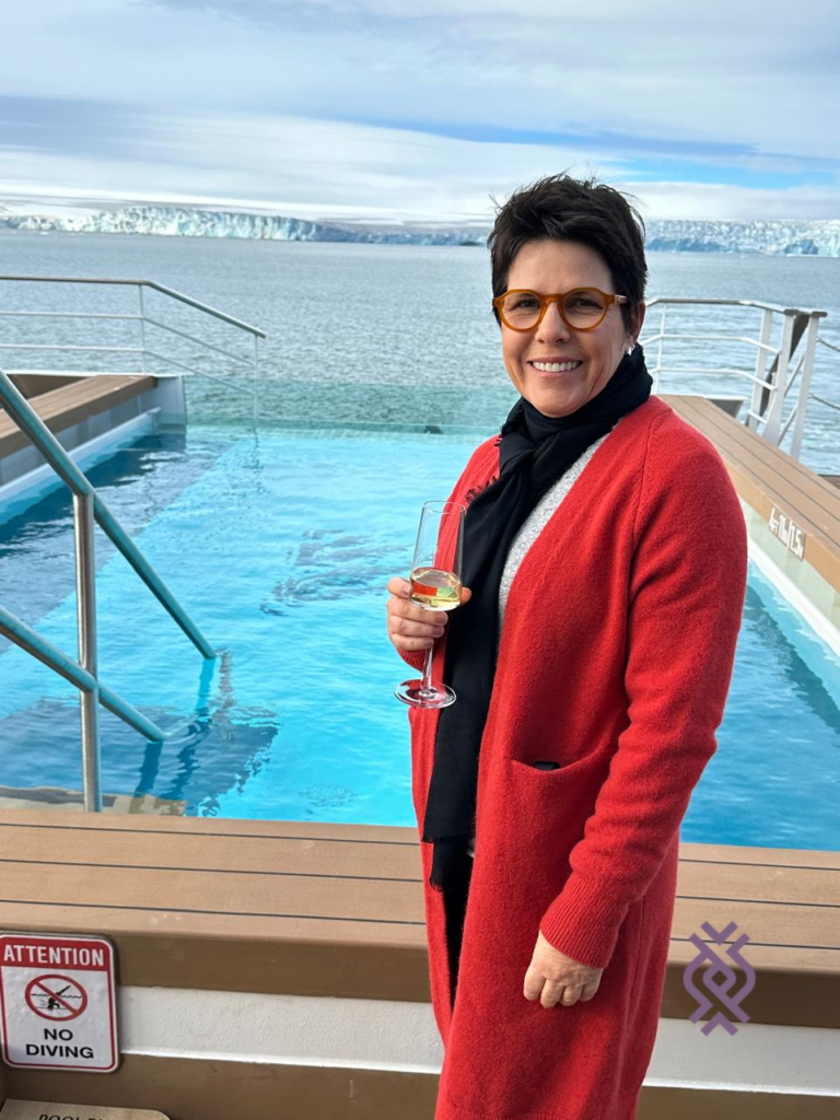 Fale com a Sabine pelo WhatsApp! Sabine Habestok, diretora operacional da Inguz Expedições, posa para foto sorrindo de costas para piscina do navio da Swan Hellenic. Ela usa um sobretudo vermelho, sorri e segura uma taça de champanhe. Ao fundo, geleiras da Antártica.
