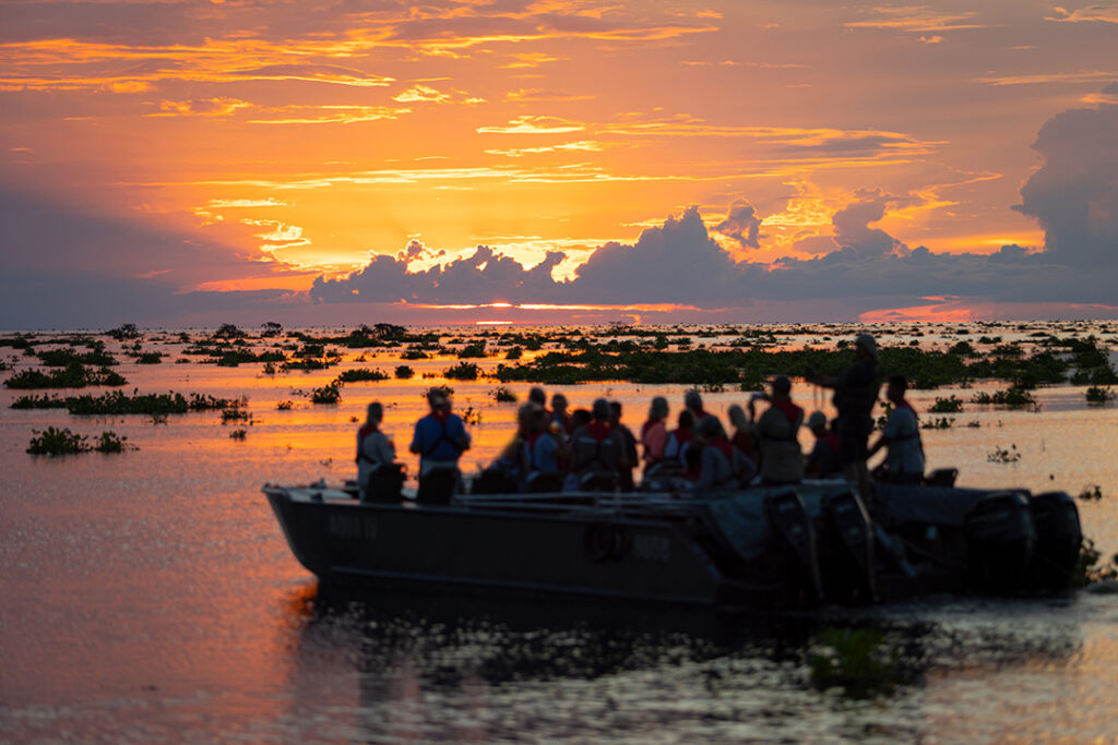 Barco a motor com diversos passageiros navegando no rio Mekong durante um pôr do sol espetacular com céu alaranjado e roxo e nuvens densas. A água reflete as cores do céu.