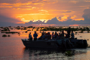 Barco a motor com diversos passageiros navegando no rio Mekong durante um pôr do sol espetacular com céu alaranjado e roxo e nuvens densas. A água reflete as cores do céu.
