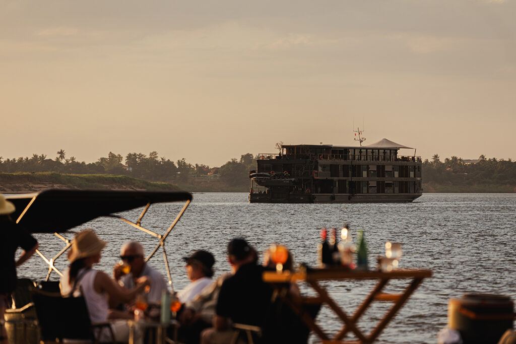 Grupo de pessoas sentadas em cadeiras de praia em primeiro plano, desfocadas, assistindo a um grande iate de luxo navegando no rio Mekong ao pôr do sol, com céu laranja e vegetação nas margens.