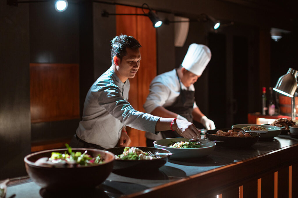 Dois chefs uniformizados em uma cozinha com iluminação indireta, servindo diversos pratos coloridos em tigelas sobre um balcão, representando a culinária da região do rio Mekong.