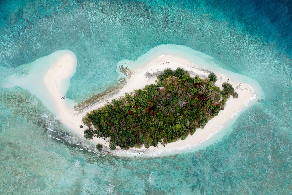 Uma foto aérea direta de uma pequena ilha tropical isolada, coberta por vegetação verde e circundada por areia branca e um vibrante recife de coral em águas azul-turquesa. A imagem representa os refúgios perfeitos que você encontra ao viajar para a Indonésia.