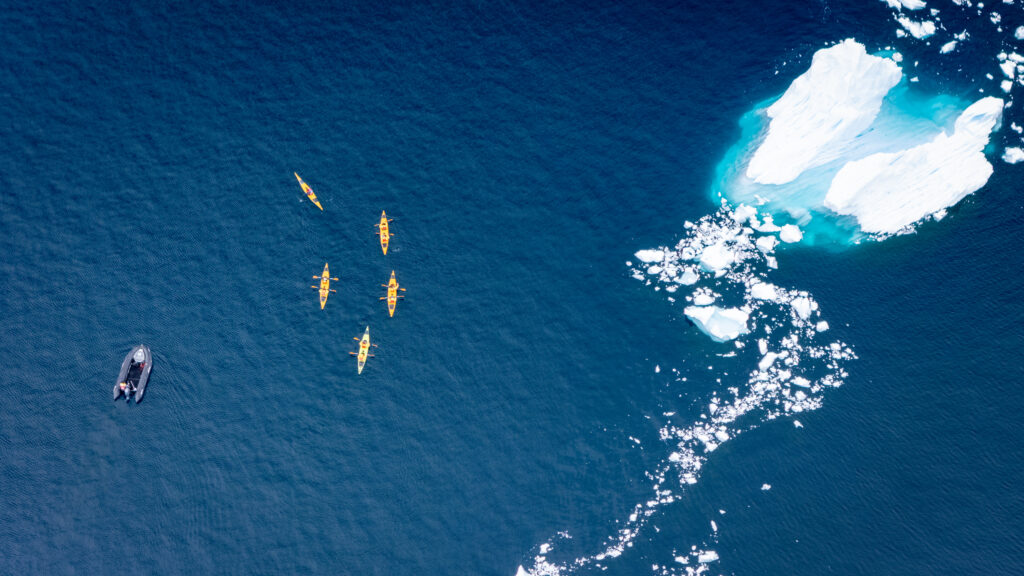 Vista aérea de caiaques amarelos navegando em águas azuis profundas ao lado de icebergs esculpidos; exploração ativa da natureza intocada no Ártico ou Antártica.