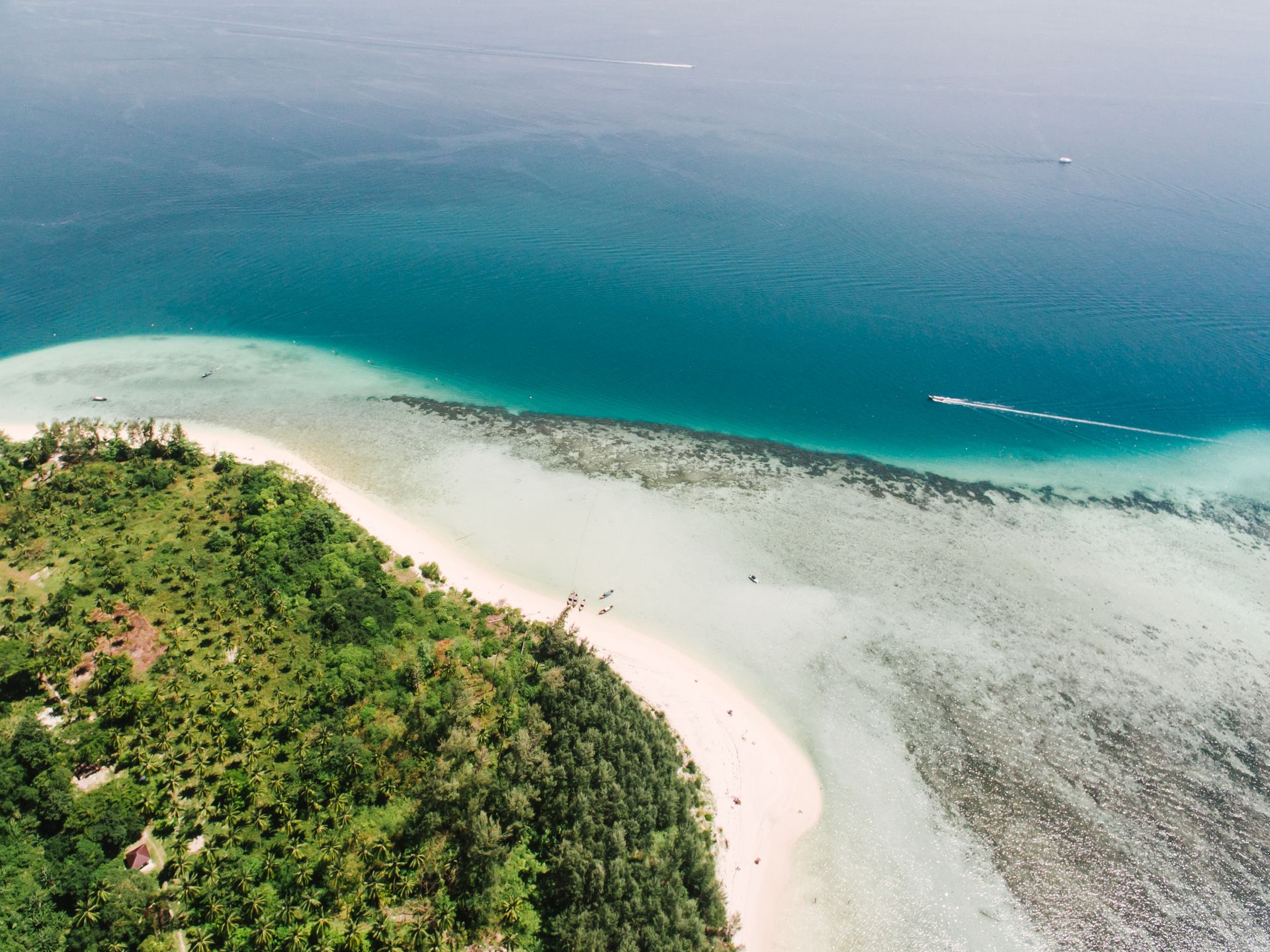 Vista aérea de uma praia paradisíaca de areia branca e águas cristalinas em tons de turquesa. A praia é contornada por uma densa vegetação tropical verde-escura. Barcos navegam calmamente pelo mar azul profundo, refletindo o sol tropical. Imagem otimizada para Inguz Expedições Marítimas.