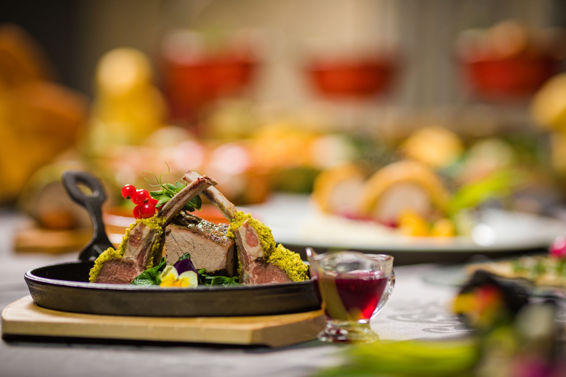 Fotografia gastronômica em close de um prato de costeletas de cordeiro assadas, com crosta de pistache verde, servidas em uma pequena frigideira de ferro fundido. O prato está guarnecido com groselhas vermelhas vibrantes e flores comestíveis sobre uma cama de vegetais verdes. Uma pequena molheira de vidro com molho de frutas vermelhas está ao lado. O fundo está em desfoque (bokeh), mostrando outros pratos de buffet e tigelas vermelhas desfocadas, indicando um evento ou restaurante requintado.