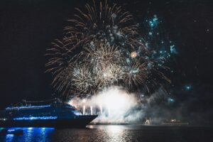 Noite em alto mar, com navio no primeiro plano e fogos de artifício ao fundo.